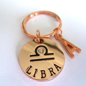 Op de afbeelding: Roségouden Weegschaal dierenriem sleutelhanger met een klein kristallen accent. De ronde bedel heeft het Weegschaal symbool en het woord "LIBRA". Een "A" bedel hangt naast de dierenriem bedel. De sleutelhanger ring is ook roségoud.