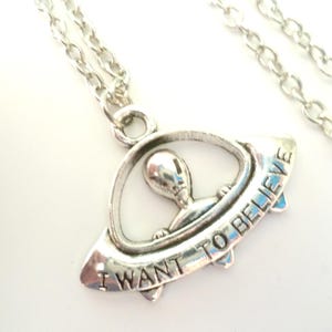 May include: Silver-toned necklace featuring a pendant with an alien figure inside a spaceship. The spaceship has the words "I WANT TO BELIEVE" engraved on it. The chain is a delicate link style.