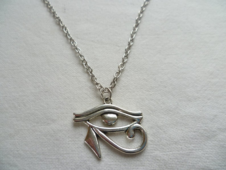 May include: Silver-toned necklace featuring an Eye of Horus pendant. The pendant is a stylized eye with intricate details, suspended from a delicate chain. The necklace is likely made of a metal alloy and is suitable for everyday wear. The design is inspired by ancient Egyptian symbolism.