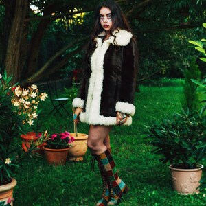 May include: A woman wearing a brown faux fur jacket with a white shearling lining, standing in a garden setting. She is wearing colorful patterned boots and has dark hair and makeup.