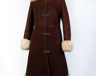 May include: A long, dark brown coat featuring a thick, cream-coloured fur collar, cuffs, and hem. The coat is secured down the front with three matching brown toggle closures. The exterior has a smooth, matte finish and long sleeves.