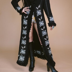 May include: A long black knit duster cardigan featuring a repeating grey and white floral pattern along the front panels and cuffs. The garment is detailed with black fringe trim along the collar, front opening, and sleeve cuffs, reaching mid-calf.