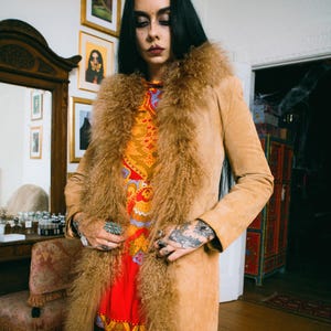 May include: A tan suede coat with a shaggy fur collar and trim. The coat is worn over a red and orange patterned dress. The image is taken indoors, with a vintage aesthetic. The coat is knee-length and has a classic silhouette.