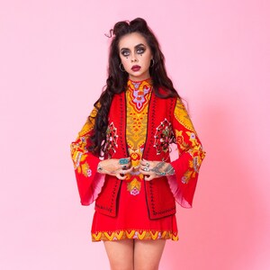 Pode incluir: Uma mulher vestindo um vestido estampado em vermelho e amarelo com um colete vermelho e botas vermelhas. Ela est&aacute; em p&eacute; em frente a um fundo rosa.