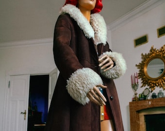 May include: A brown suede coat with white faux fur trim on the collar, cuffs, and hem. The coat is worn by a mannequin with red hair and sunglasses. The mannequin is also wearing red boots.