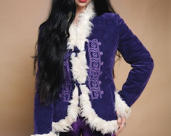 Könnte beinhalten: Eine violette Samtjacke mit weißem Kunstfellbesatz und gestickten Details. Die Jacke wird mit einer passenden lila Samthose kombiniert. Das Outfit hat eine Vintage- und Bohème-Ästhetik.