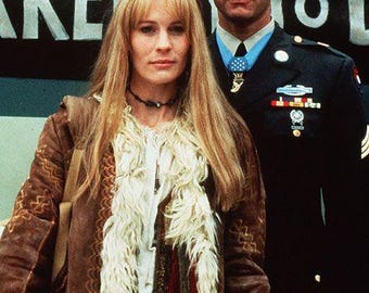 Puede incluir: Una persona con cabello rubio largo viste una chaqueta de gamuza marrón con ribete de piel sintética blanca y una gargantilla negra, junto a una persona con uniforme militar azul oscuro con gorra, medallas y cintas, frente a un letrero negro.