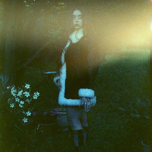 May include: A woman wearing a black dress with a white fur trim stands in a grassy area. The background is blurry and out of focus.