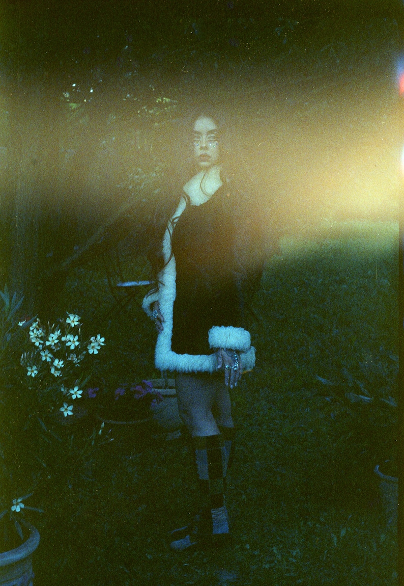 May include: A woman wearing a black dress with a white fur trim stands in a grassy area. The background is blurry and out of focus.
