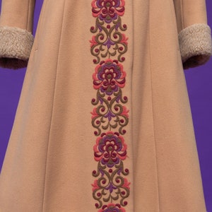May include: A tan coat with a long, embroidered floral design down the front. The embroidery is in shades of pink, purple, and brown. The coat has a fur trim around the cuffs and hem.
