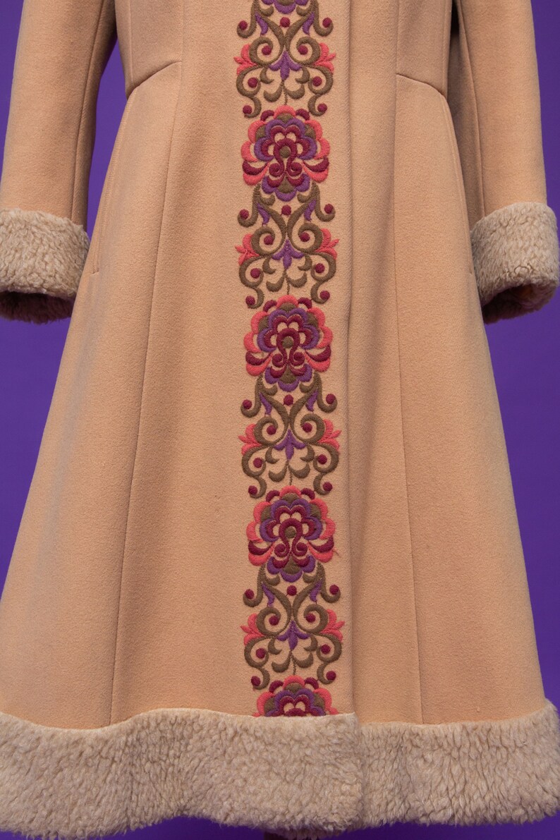 May include: A tan coat with a long, embroidered floral design down the front. The embroidery is in shades of pink, purple, and brown. The coat has a fur trim around the cuffs and hem.