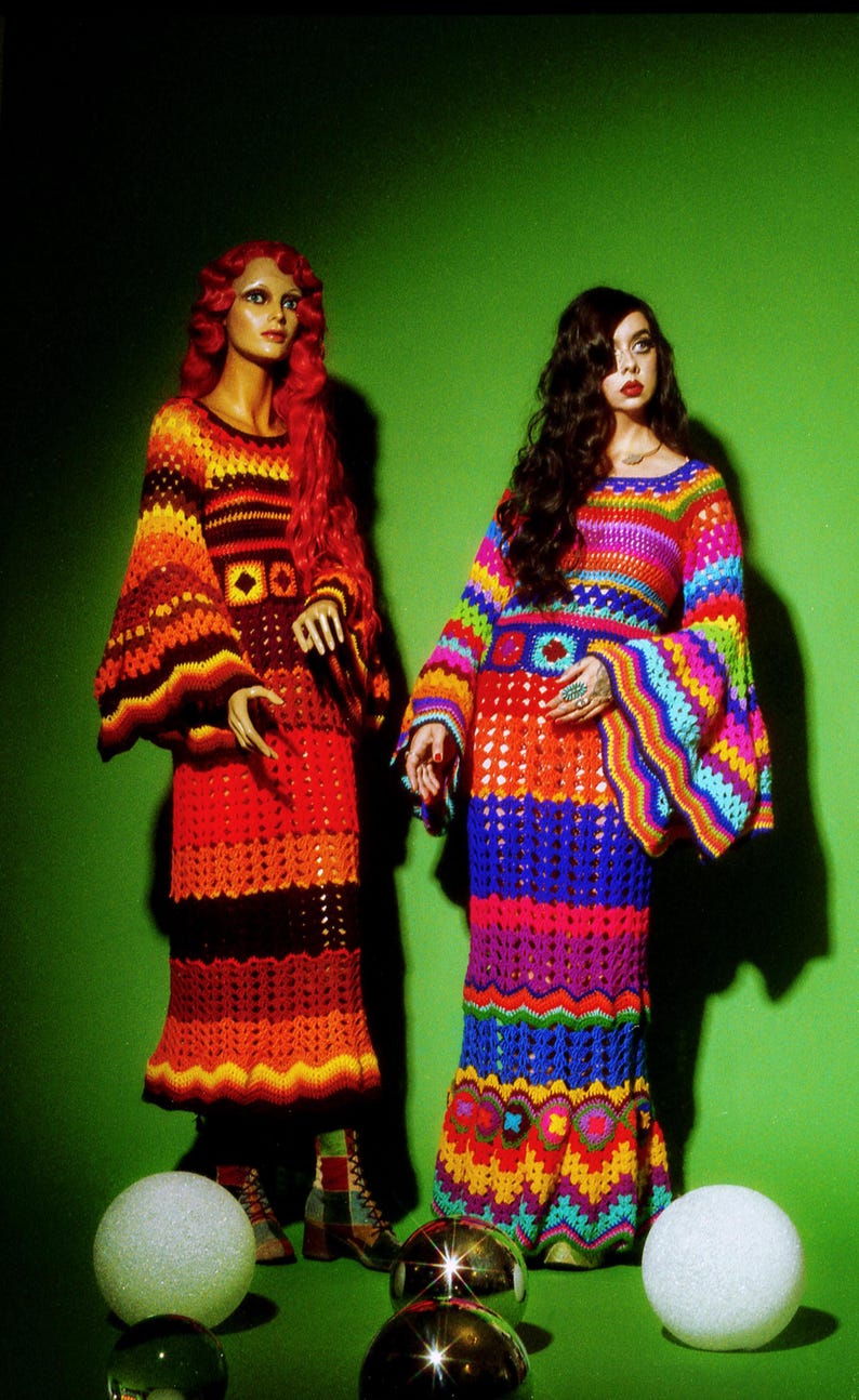 May include: Two mannequins wearing colorful, crochet dresses with bell sleeves and flared skirts. One dress is in shades of red, orange, and brown. The other is a rainbow of colors. The background is a solid green.