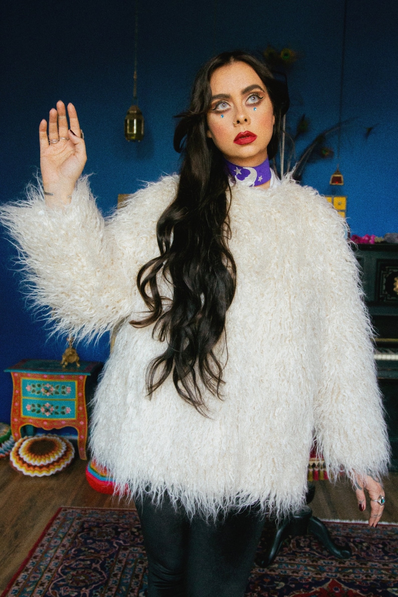 May include: A woman with long black hair wearing a white fluffy jacket and black pants. She is standing in a room with a blue wall and a rug on the floor. She has blue eyeshadow and red lipstick.