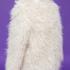 May include: A white faux fur jacket with a long, shaggy pile. The jacket is on a mannequin and is displayed against a purple background.