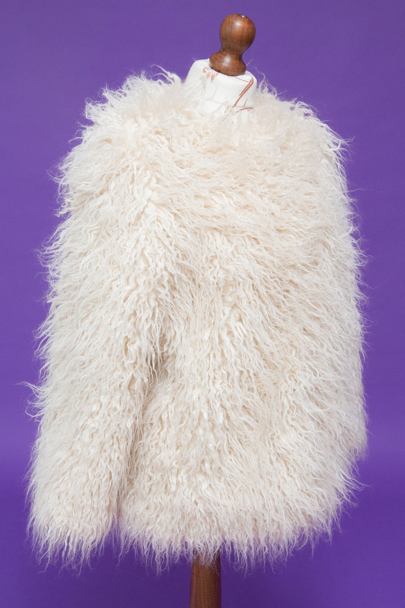May include: A white faux fur jacket with a long, shaggy pile. The jacket is on a mannequin and is displayed against a purple background.