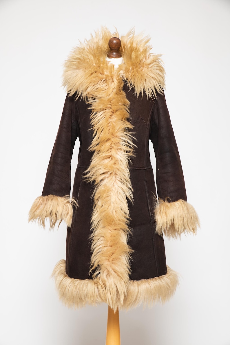 Authentic vintage Penny Lane coat in genuine sheepskin with shearling fur trim. Women's 70s Afghan coat inspired by Woodstock fashion and classic boho winter style..May include: A dark brown coat with a tan faux fur collar, trim, and cuffs. The long coat is displayed on a mannequin and has a vintage, retro style. The coat is knee-length and has a button closure.