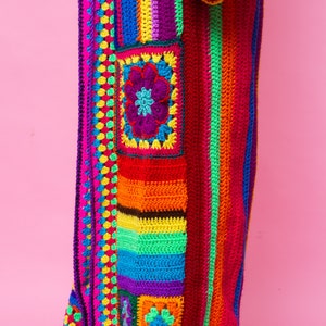 K&ouml;nnte beinhalten: Ein lebendiger, handgefertigter H&auml;kelmantel im Patchwork-Design. Der Mantel weist ein Farbspektrum auf, darunter Rosa, Rot, Orange, Gelb, Gr&uuml;n, Blau und Lila. Er hat ein Blumenmotiv und ein Streifenmuster, mit einer langen Silhouette.