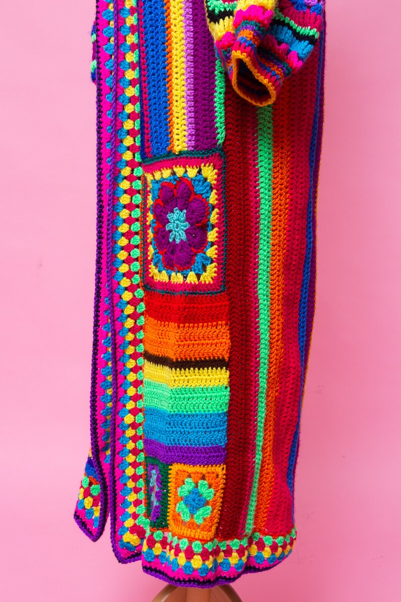K&ouml;nnte beinhalten: Ein lebendiger, handgefertigter H&auml;kelmantel im Patchwork-Design. Der Mantel weist ein Farbspektrum auf, darunter Rosa, Rot, Orange, Gelb, Gr&uuml;n, Blau und Lila. Er hat ein Blumenmotiv und ein Streifenmuster, mit einer langen Silhouette.
