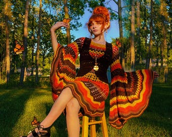 May include: A woman wearing a colourful crochet dress with bell sleeves sits on a wooden stool in a woodland setting.  She is surrounded by monarch butterflies.