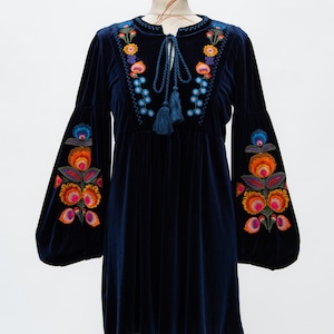 May include: A navy blue velvet dress with long, bell sleeves. The dress features colourful floral embroidery on the neckline and sleeves. A tie with tassels adorns the neckline. The dress is displayed on a mannequin.