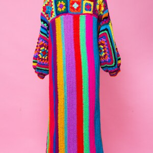 K&ouml;nnte beinhalten: Ein langer, handgefertigter H&auml;kelmantel in leuchtenden Farben. Der Mantel hat ein Patchwork-Design an Schultern und &Auml;rmeln, mit einem Streifenmuster am K&ouml;rper. Die Farben sind Rot, Orange, Gelb, Gr&uuml;n, Blau und Lila. Der Mantel wird auf einer Schaufensterpuppe pr&auml;sentiert.