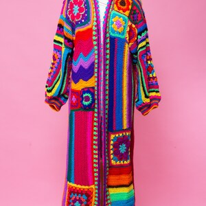 K&ouml;nnte beinhalten: Ein langer, farbenfroher H&auml;kelmantel, der auf einer Schaufensterpuppe pr&auml;sentiert wird. Der Mantel hat ein Patchwork-Design mit Quadraten und Streifen in einem Regenbogen von Farben, darunter Rot, Rosa, Gelb, Gr&uuml;n, Blau und Lila. Die &Auml;rmel sind lang und der Mantel ist vorne offen.