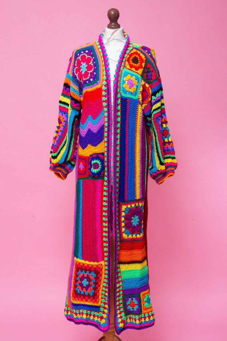 K&ouml;nnte beinhalten: Ein langer, farbenfroher H&auml;kelmantel, der auf einer Schaufensterpuppe pr&auml;sentiert wird. Der Mantel hat ein Patchwork-Design mit Quadraten und Streifen in einem Regenbogen von Farben, darunter Rot, Rosa, Gelb, Gr&uuml;n, Blau und Lila. Die &Auml;rmel sind lang und der Mantel ist vorne offen.
