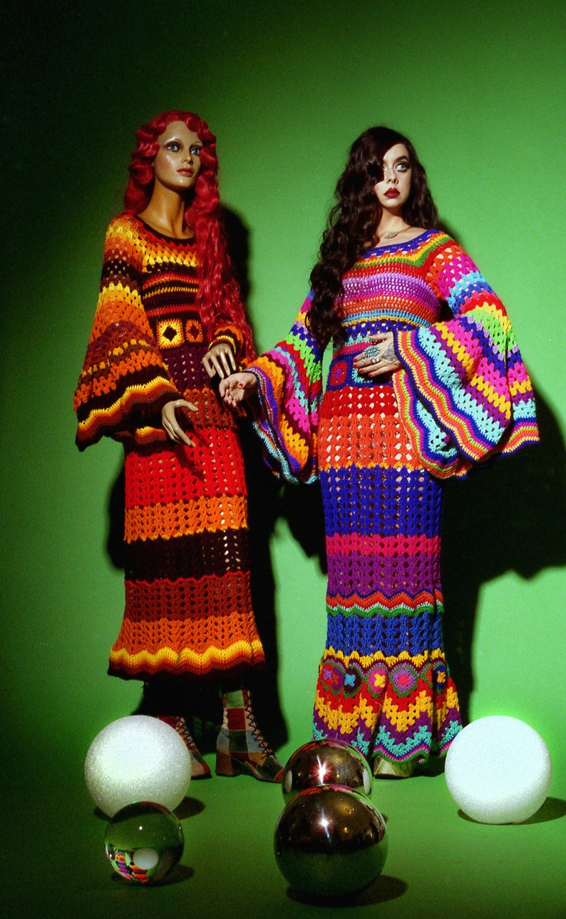 May include: Two mannequins display colorful, handmade crochet dresses against a green backdrop. One dress features orange, brown, and yellow hues, while the other showcases a rainbow of colors. Decorative spheres are in the foreground.