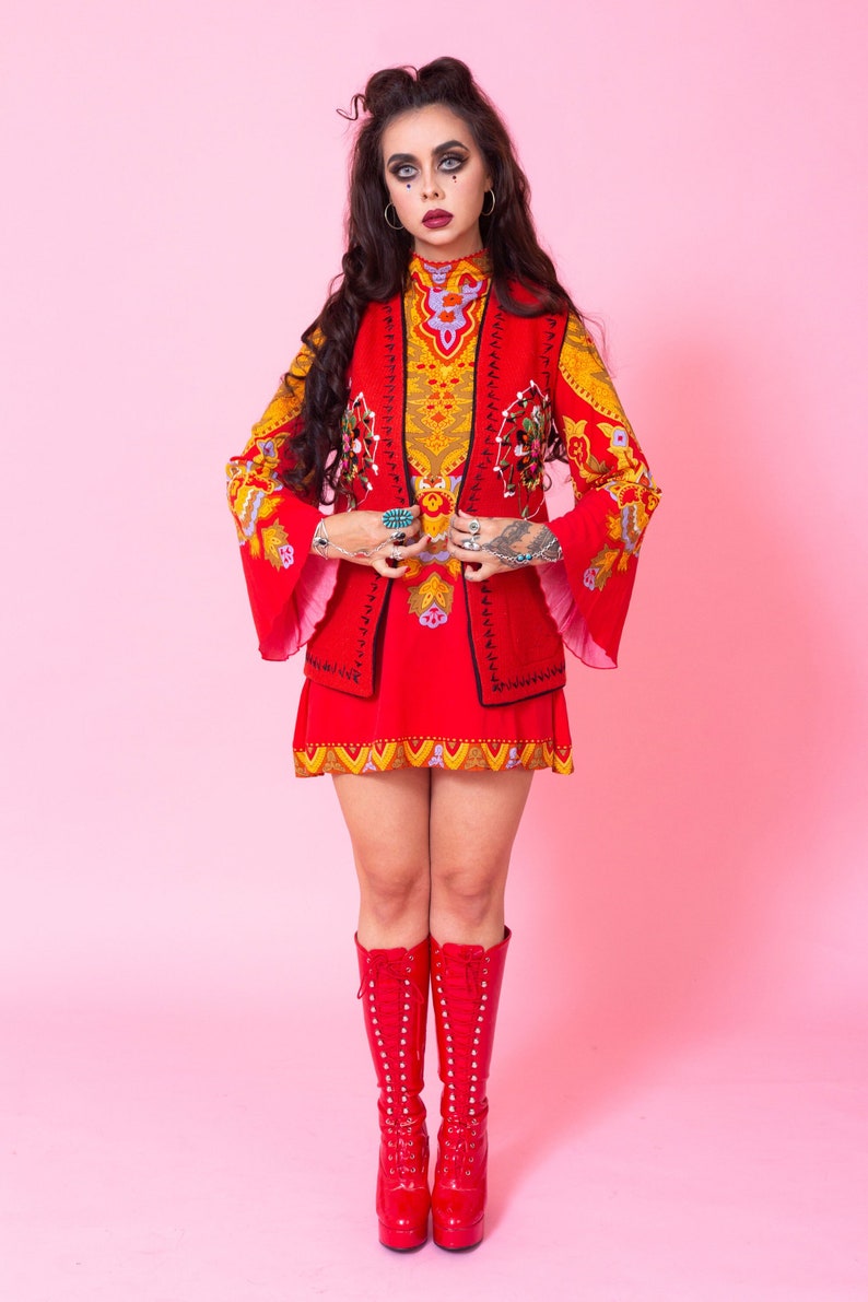 Pode incluir: Uma mulher vestindo um vestido estampado em vermelho e amarelo com um colete vermelho e botas vermelhas. Ela est&aacute; em p&eacute; em frente a um fundo rosa.