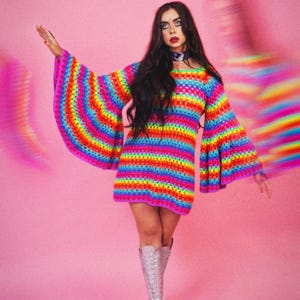 Könnte beinhalten: Eine Frau trägt ein regenbogenfarbenes gestricktes Kleid mit langen Glockenärmeln und silbernen Plateau-Stiefeln. Sie steht vor einem rosa Hintergrund.