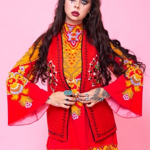 Pode incluir: Uma mulher vestindo um vestido estampado vermelho e amarelo com um colete vermelho. O colete tem bordados pretos e um design floral. A mulher tem cabelo escuro e est&aacute; usando maquiagem.