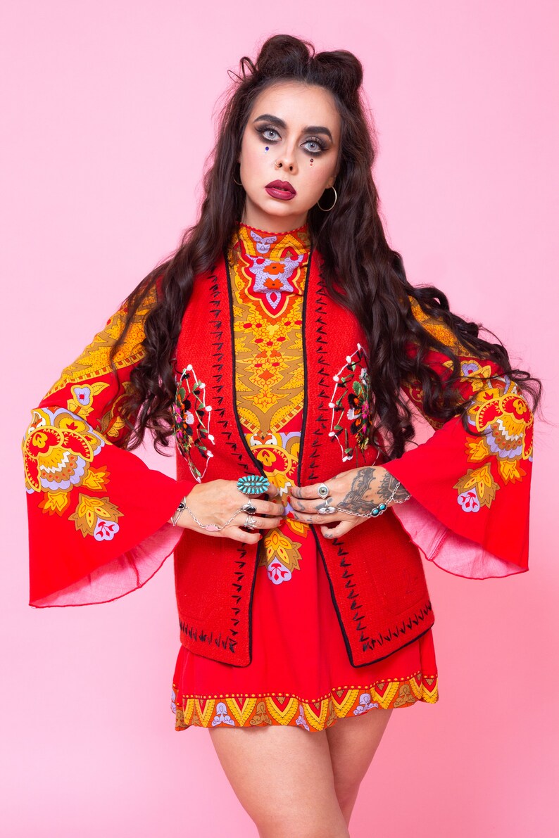 Pode incluir: Uma mulher vestindo um vestido estampado vermelho e amarelo com um colete vermelho. O colete tem bordados pretos e um design floral. A mulher tem cabelo escuro e est&aacute; usando maquiagem.