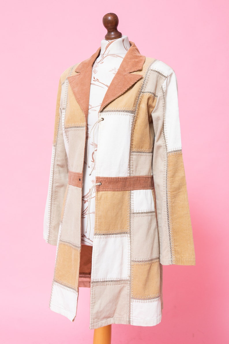 K&ouml;nnte beinhalten: Eine lange Patchworkjacke in Braun, Beige und Wei&szlig;. Die Jacke hat einen Kragen und einen Knopfverschluss. Sie ist aus Cord und hat ein gestepptes Design.