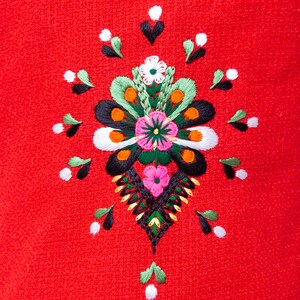 Pode incluir: Um fundo de tecido vermelho com um desenho floral bordado colorido. O desenho apresenta uma flor central com p&eacute;talas rosa, brancas e verdes, cercada por flores e folhas menores em tons de verde, preto, laranja e branco.