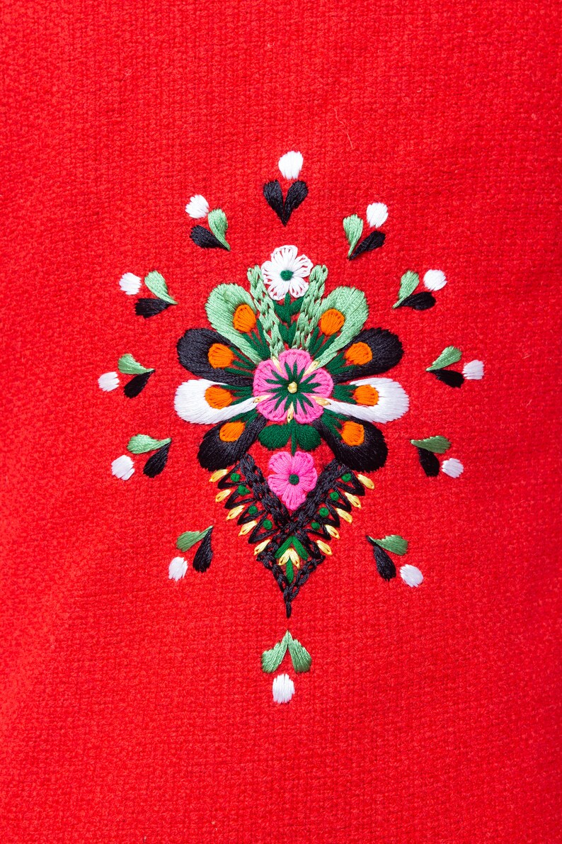 Pode incluir: Um fundo de tecido vermelho com um desenho floral bordado colorido. O desenho apresenta uma flor central com p&eacute;talas rosa, brancas e verdes, cercada por flores e folhas menores em tons de verde, preto, laranja e branco.