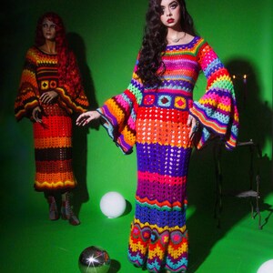 May include: A vibrant, handmade crochet dress with bell sleeves and a flared skirt, featuring a rainbow of colors including red, orange, yellow, green, blue, and purple. A mannequin in a similar dress is in the background. The dresses are on a green background.