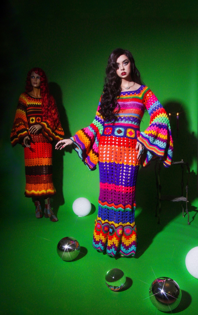 May include: A vibrant, handmade crochet dress with bell sleeves and a flared skirt, featuring a rainbow of colors including red, orange, yellow, green, blue, and purple. A mannequin in a similar dress is in the background. The dresses are on a green background.