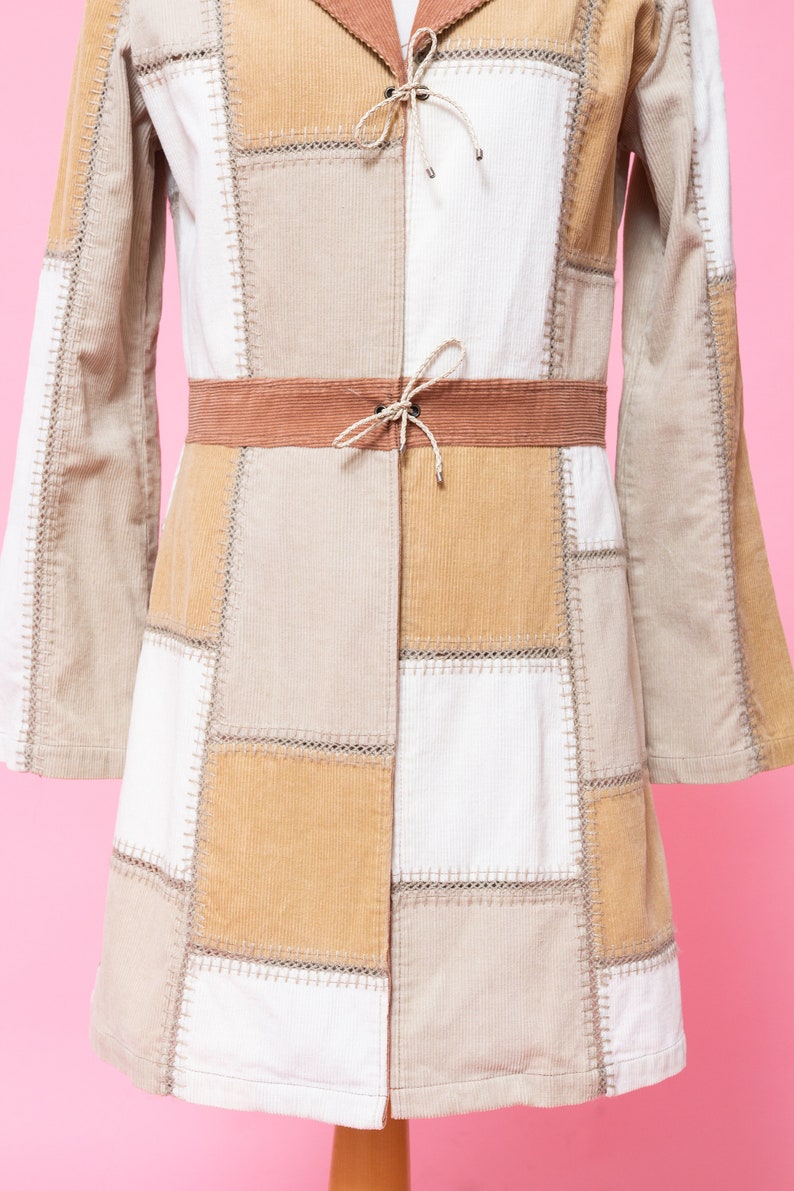 K&ouml;nnte beinhalten: Eine lang&auml;rmelige Patchwork-Jacke mit braunem Cordkragen und G&uuml;rtel. Die Jacke besteht aus verschiedenen Farbt&ouml;nen von beige und wei&szlig;em Cordstoff.