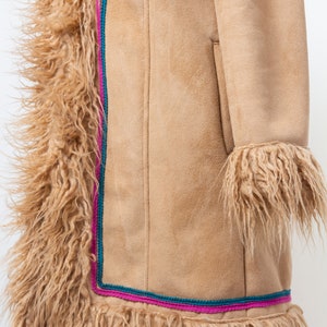 May include: A tan suede coat with a long, fuzzy trim around the edges and sleeves. The trim is a light brown color and the coat has a pink and teal trim along the bottom.