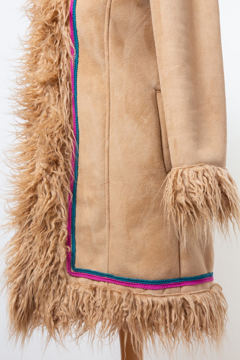 May include: A tan suede coat with a long, fuzzy trim around the edges and sleeves. The trim is a light brown color and the coat has a pink and teal trim along the bottom.