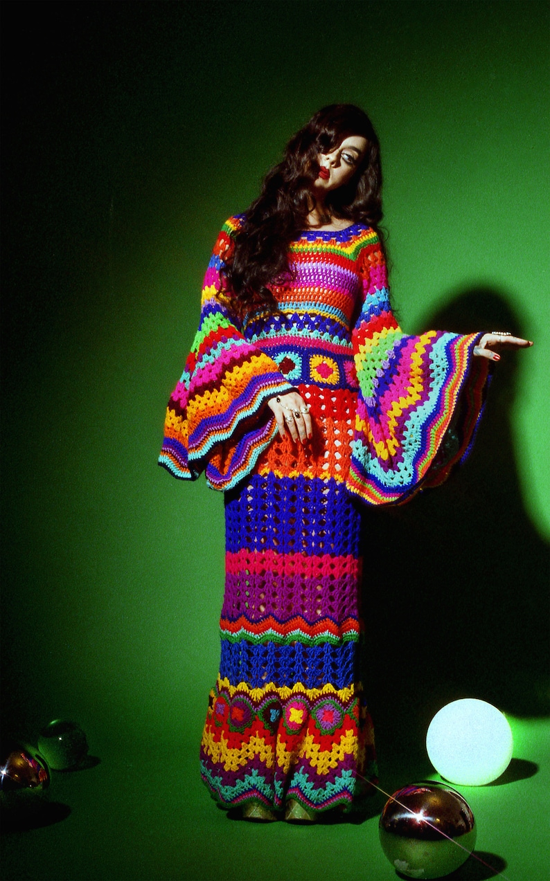 May include: A vibrant, handmade crochet dress in a rainbow of colors. The dress features long, flared sleeves and a fitted bodice that flows into a floor-length skirt. The intricate crochet work showcases a variety of patterns and textures, creating a bohemian aesthetic.