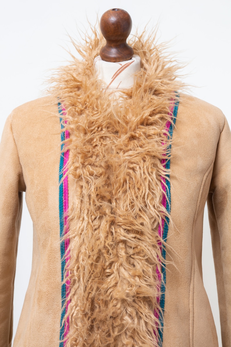 May include: A tan suede jacket with a long, fluffy brown fur collar. The jacket has a vertical stripe down the front, with pink, teal, and green accents.