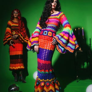 May include: A woman wearing a vibrant, handmade crochet dress with long, flared sleeves. The dress features a rainbow of colors, including red, orange, yellow, green, blue, and purple. A mannequin in a similar dress stands nearby.