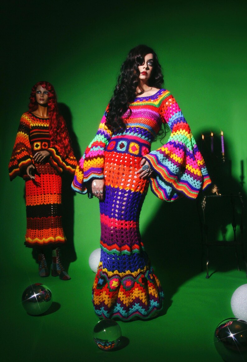 May include: A woman wearing a vibrant, handmade crochet dress with long, flared sleeves. The dress features a rainbow of colors, including red, orange, yellow, green, blue, and purple. A mannequin in a similar dress stands nearby.
