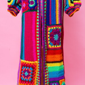 K&ouml;nnte beinhalten: Ein lebendiger, handgefertigter H&auml;kelcardigan in einem Patchwork-Design. Das lange, offen geschnittene Kleidungsst&uuml;ck hat einen Farbenrausch, darunter Rosa, Lila, Gelb, Gr&uuml;n und Blau. Die &Auml;rmel sind puffig und die Kanten sind mit einer Kontrastfarbe versehen.