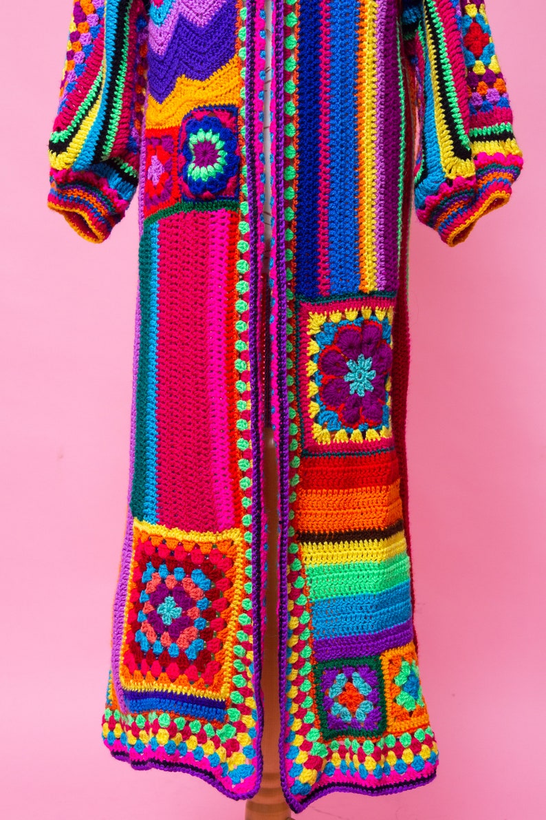 K&ouml;nnte beinhalten: Ein lebendiger, handgefertigter H&auml;kelcardigan in einem Patchwork-Design. Das lange, offen geschnittene Kleidungsst&uuml;ck hat einen Farbenrausch, darunter Rosa, Lila, Gelb, Gr&uuml;n und Blau. Die &Auml;rmel sind puffig und die Kanten sind mit einer Kontrastfarbe versehen.