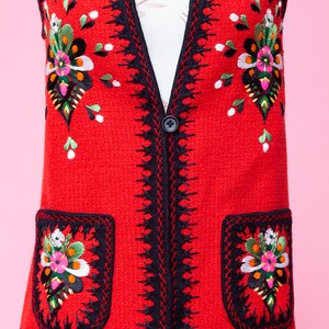 Pode incluir: Um colete vermelho sem mangas com guarni&ccedil;&otilde;es pretas e bordados florais coloridos. O colete tem dois bolsos com flores bordadas na frente.