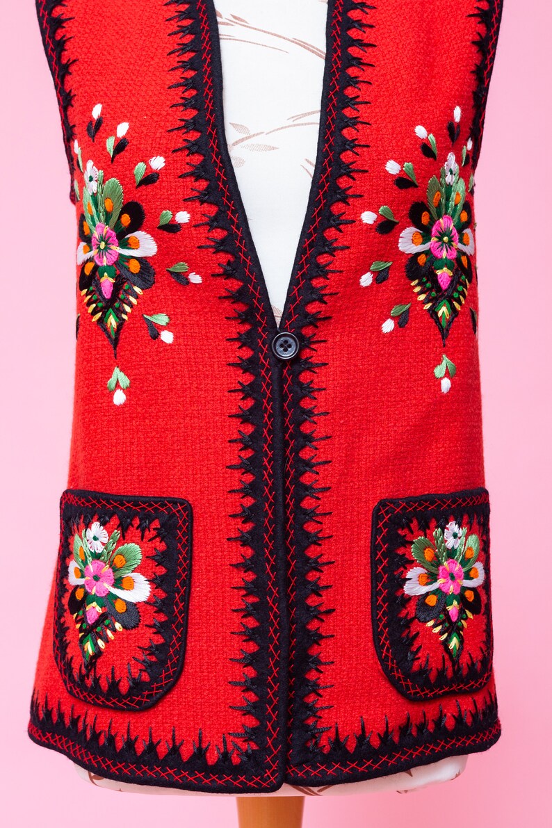 Pode incluir: Um colete vermelho sem mangas com guarni&ccedil;&otilde;es pretas e bordados florais coloridos. O colete tem dois bolsos com flores bordadas na frente.
