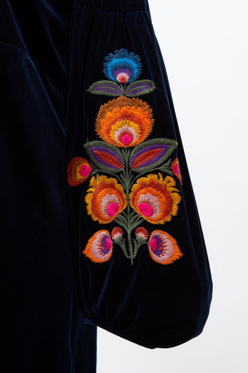 May include: Dark navy blue velvet fabric with a floral embroidery design. The embroidery features orange, pink, purple, and blue flowers with green leaves. The sleeve is gathered at the wrist.