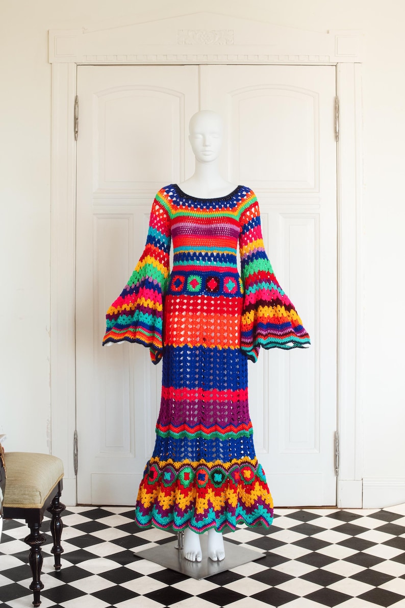 May include: A vibrant, handmade crochet dress in a rainbow of colors, including red, orange, blue, and green. The dress features a round neckline, long bell sleeves, and a flared skirt. It is displayed on a mannequin.